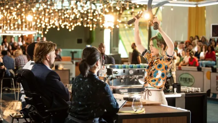 Coffee Wars: A thirty-something White woman with green hair holds aloft two swords during a coffee-making competition