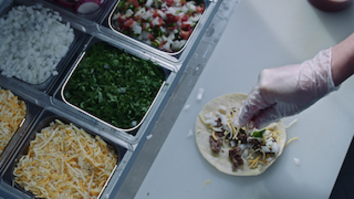 Still from Poisoned: The Dirty Truth About Your Food: A taco on a white counter with various ingredients being added by an individual, wearing a clear glove on their right hand