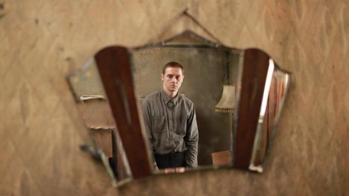 Possum: The reflection of a White man with ginger hair is shown as he looks into a mirror hanging on a fading ochre-colored wall