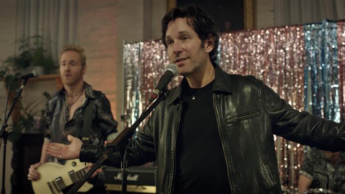 Power Ballad movie: Wedding singer Rick (Paul Rudd) holds his arms out as he talks into a mic while his band stands behind him on a low-budget stage