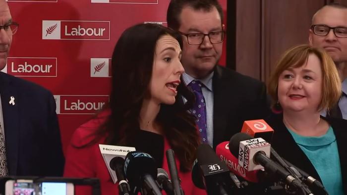 Prime Minister movie: Jacinda Ardern addresses her nation of New Zealand at a press conference, White New Zealander men and women in suits behind her