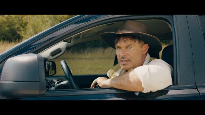 A man wearing an adventure hat sits in a black car with his arm propped outside the window. He is looking at something intently in the distance. 