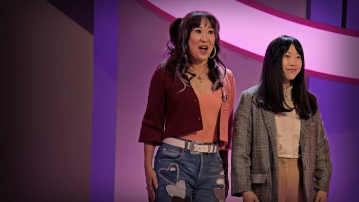 Quiz Lady movie: Sandra Oh wears pink and red top, blue jeans, and hoop earrings center stands next to Awkwafina in tweed sports coat on right