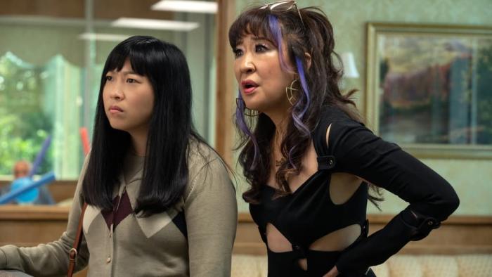 Quiz Lady movie: Awkwafina in argyle tan sweater top right stands next to Sandra Oh in black cut out dress on right