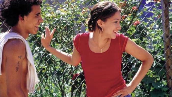 Raising Victor Vargas movie: Two young people laughing together outdoors near a chain-link fence with greenery in the background.