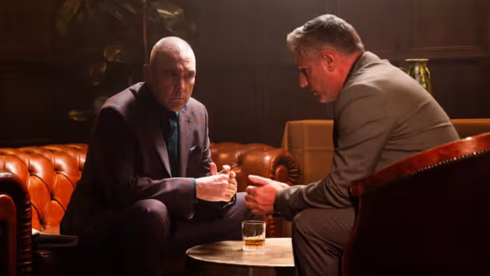 Reckless: Two men in suits having a serious conversation on a leather couch with a drink on the table