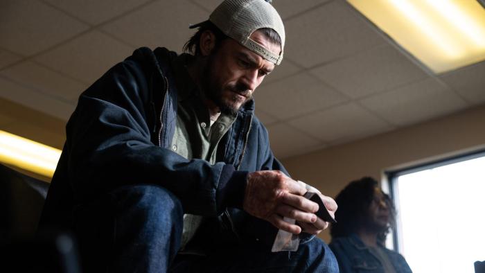 Red Right Hand Movie: Wearing a coat and a backward baseball cap, Cash (Orlando Bloom) sits in a chair and examines something he holds in his hands, his face deep in concentration
