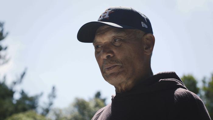 Still from Reggie: African American and retired baseball player Reggie Jackson is looking at his surrounding area, wearing a dark brown Houston Astros baseball cap with a capital white letter H and a dark brown hoodie. Jackson is special advisor the organization and team's owner Jim Crane 