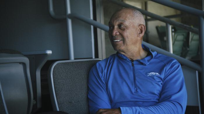 Still from Reggie: African American and retired baseball outfielder Reggie Jackson seated in a stadium and chatting with and looking toward former baseball colleagues, shown off camera, wearing an an electric blue activewear long-sleeved mock turtleneck top with a black zipper and small white logo