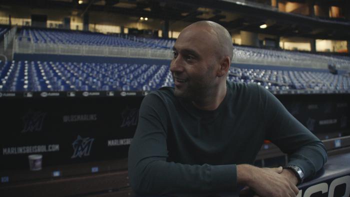 Still from Reggie: Retired Yankees shortstop Derek Jeter standing in stadium bleachers chatting with and looking toward former baseball colleague Reggie Jackson, shown off camera, wearing a dark green, long-sleeved activewear top 