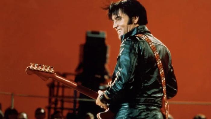 Return of the King: Elvis Presley stands smiling over his shoulder against a dark orange backdrop holding a guitar and wearing a black leather suit.