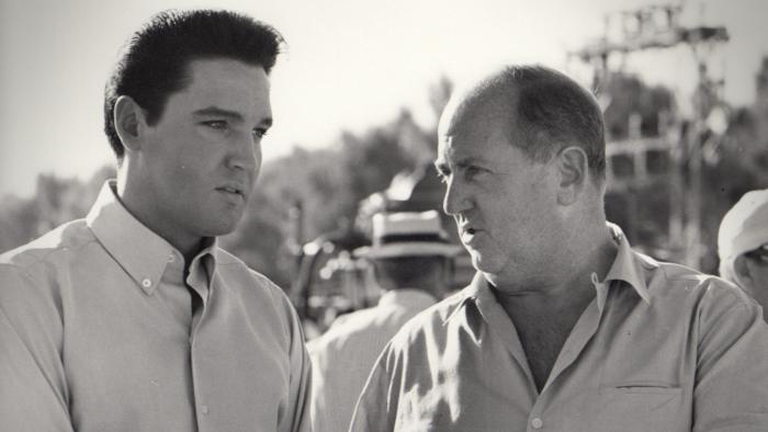 Return of the King: A black and white still of Elvis standing outdoors talking intently with his manager, Colonel Tom Parker.
