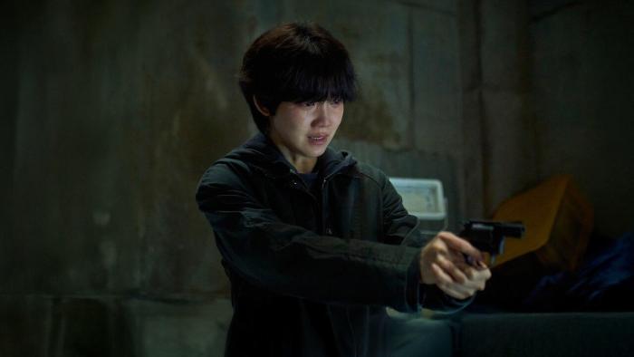Revelations movie: Korean woman with short hair in black jacket center holds gun forward in dark warehouse