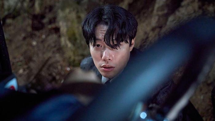 Revelations movie: Through foreground of car steering wheel, a Korean man with short black hair looks up, muddy terrain behind him