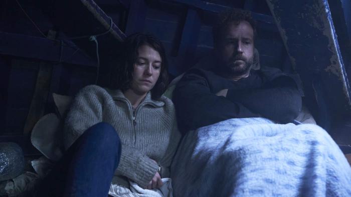 Rich Flu: A man and woman sitting close together under a blanket, appearing thoughtful and somber