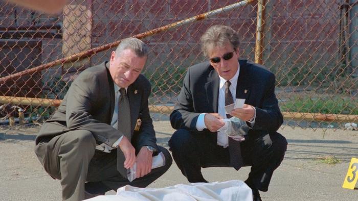 Robert De Niro and Al Pacino crouch down at a crime scene in suits, examining the area.
