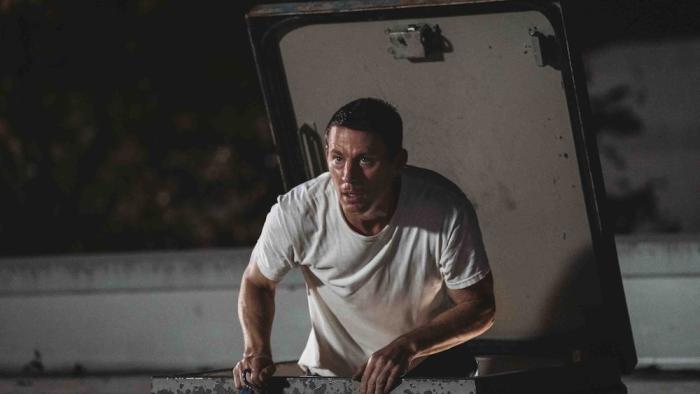 Roofman Movie: Jeffrey (Channing Tatum) climbs out of a trap door on a roof, looking shocked