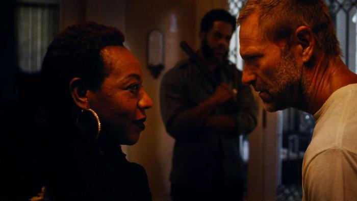 Rumble Through the Dark Movie: Momma Sweet (Marianne Jean-Baptiste, left) confronts Jack "The Butcher" Boucher (Aaron Eckhart, right), while an armed man stands, out of focus, in the background