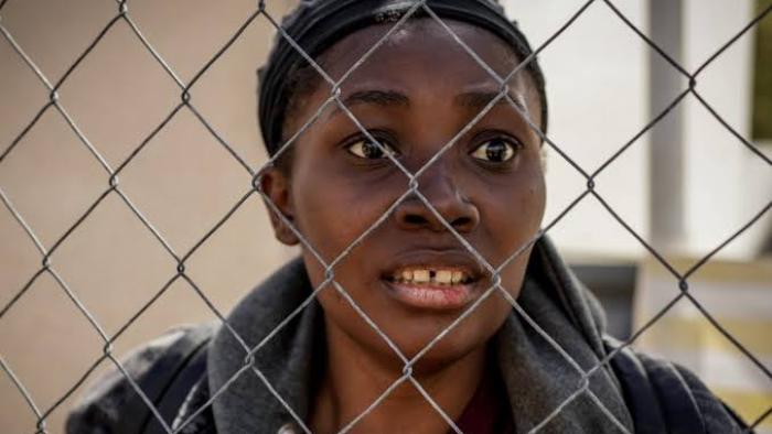 Run: A Black woman looking through a chain-link fence, appearing worried