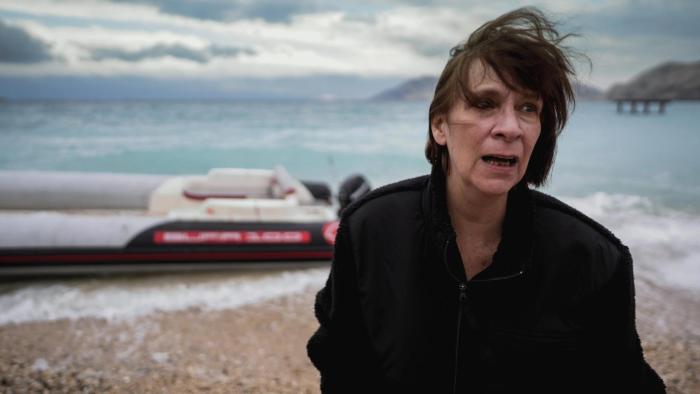 Run: Windblown White woman stands on beach, cloudy sky, boat and sea in background