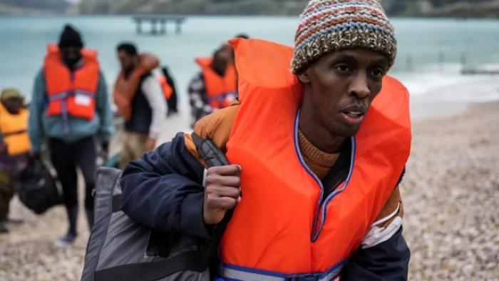 Run: Group wearing life jackets walks on a pebble beach, carrying bags