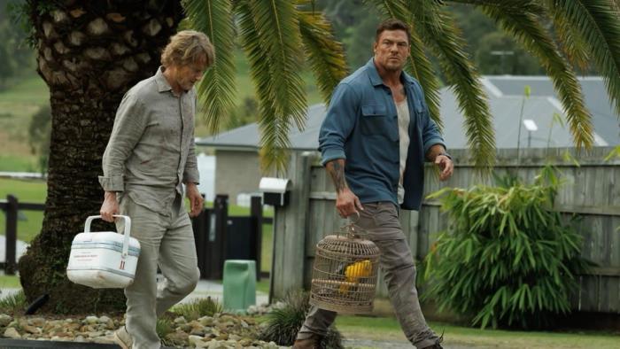 Runner movie: Alan Ritchson, holding a birdcage, walks next to Owen Wilson, holding a cooler