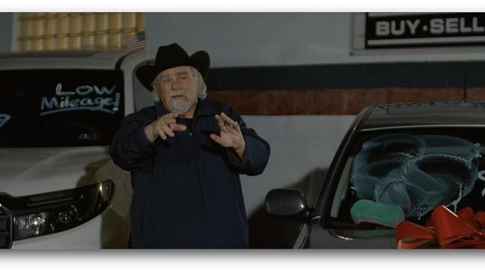 Saint Nick of Bethlehem movie: Duane Whitaker gestures with his hands as he stands next to cars in his car lot. 