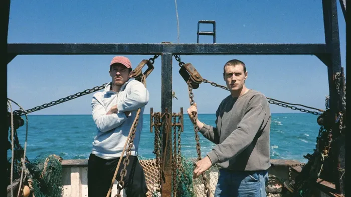 Rose of Nevada: Two men stand on a boat with ocean in the background under a clear blue sky