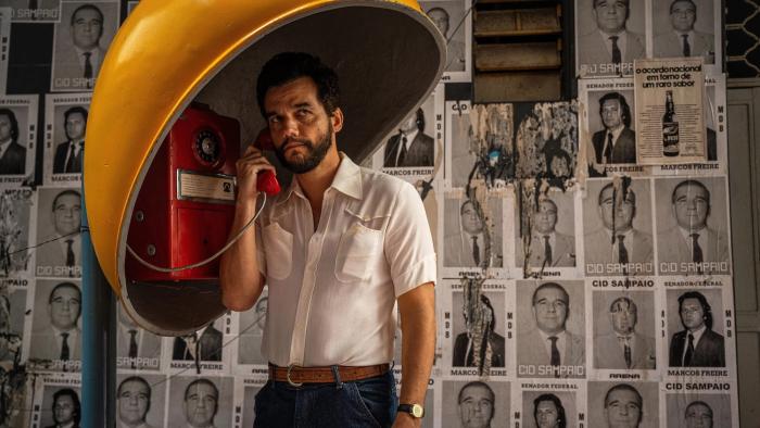 The Secret Agent movie: Wagner Moura stands holding a receiver to his ear in a yellow phone booth with political posters in the background