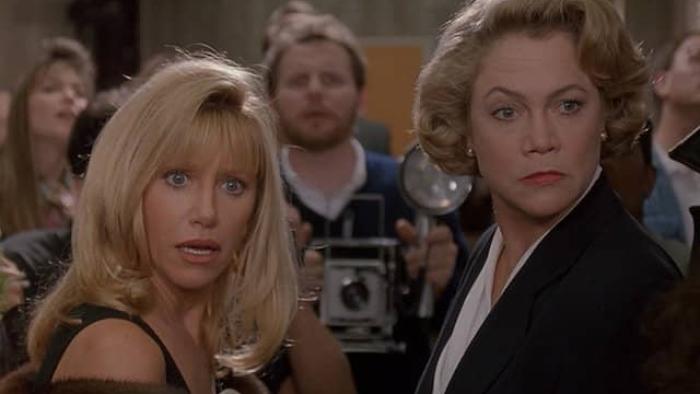 Serial Mom Movie: Two blond women look towards the camera in a crowd, one wearing a fur coat and the other a suit