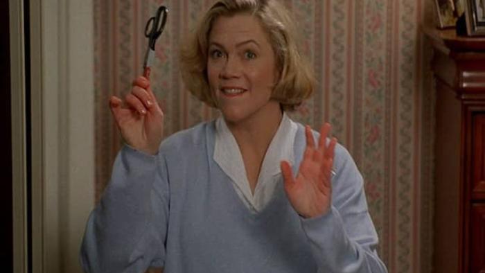 Serial Mom Movie: A mom cheekily holds up a pair of scissors inside a house with patterned wallpaper