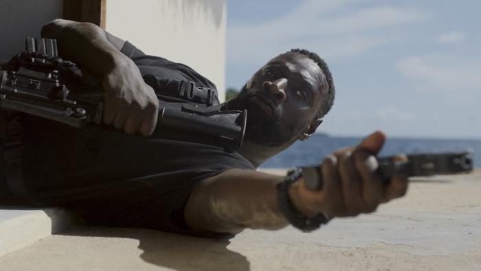 Shadow Force Movie: Isaac (Omar Sy) lies on his back in an open doorway and aims his pistol at an attacking foe