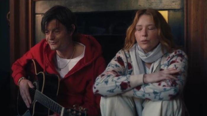 She Is Love: A White man sits against the living room wall playing guitar, with a White woman sitting next to him