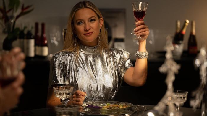 Shell Movie: In a silver dress, Zoe (Kate Hudson) raises a glass of wine at what appears to be a fancy dinner party