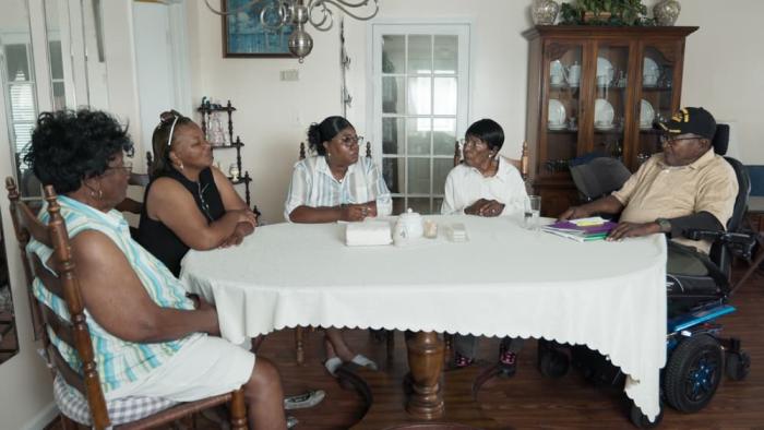 Silver Dollar Road movie: Four Black women and one Black man in motorized chair sit around white dining table in dining room of house