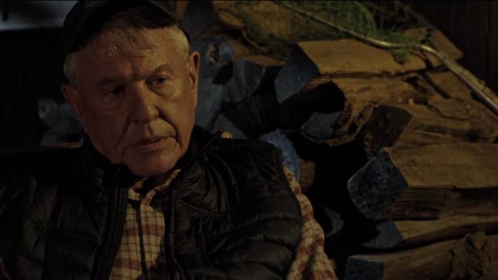 Sniper: Assassin's End: Tom Berenger sits and talks in front of wood pile.