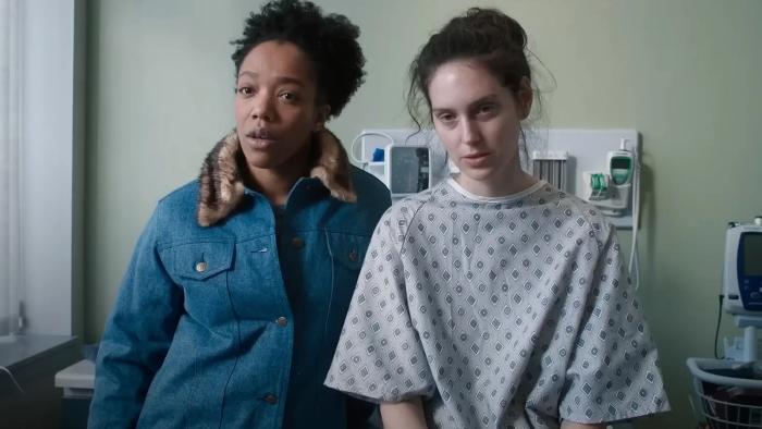 Sorry, Baby movie: Lydie (Naomi Ackie) stands next to Agnes (Eva Victor), who's sitting on a medical table wearing a hospital gown