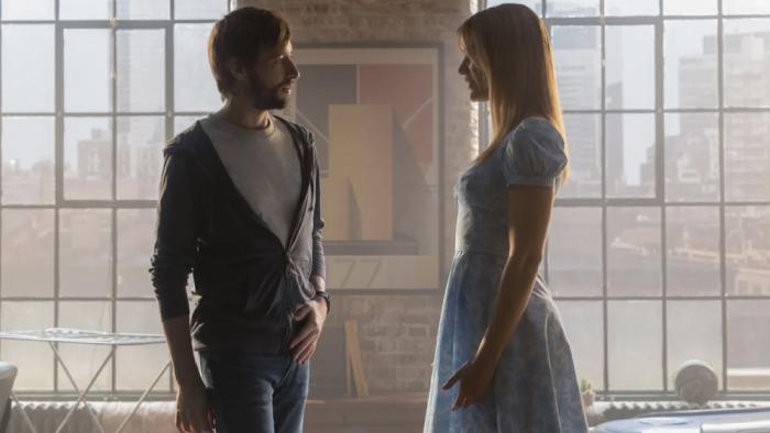 Soulm8te movie: Standing in front of picture windows, David, in a hoodie, speaks to the robot Sara, who's wearing a blue dress