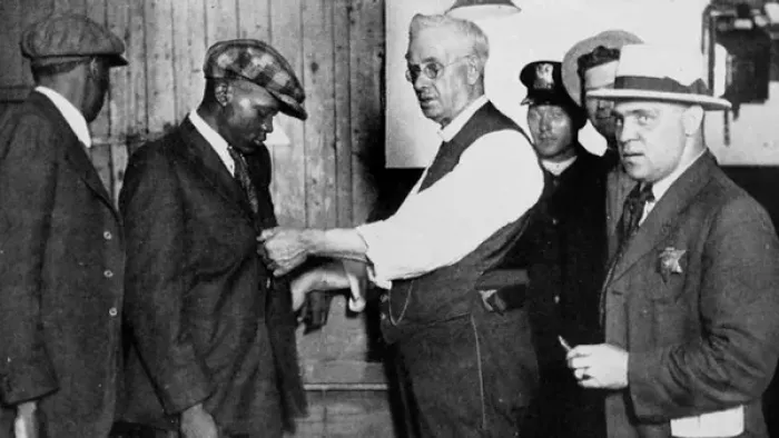 Still from Sound of the Police: A White police officer (right) searches a Black man for firearms at a Chicago police station in 1922, while surrounded by other law enforcement officials, with individuals wearing business and police attire that includes caps, white shirts, and dark pants