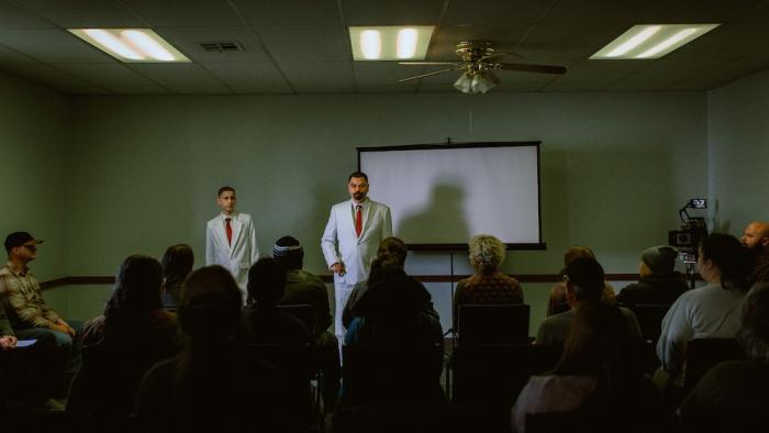 Sovereign Movie: White-suited Jerry Kane speaks to a crowd in a conference room, his nearby son, Joe, dressed alike