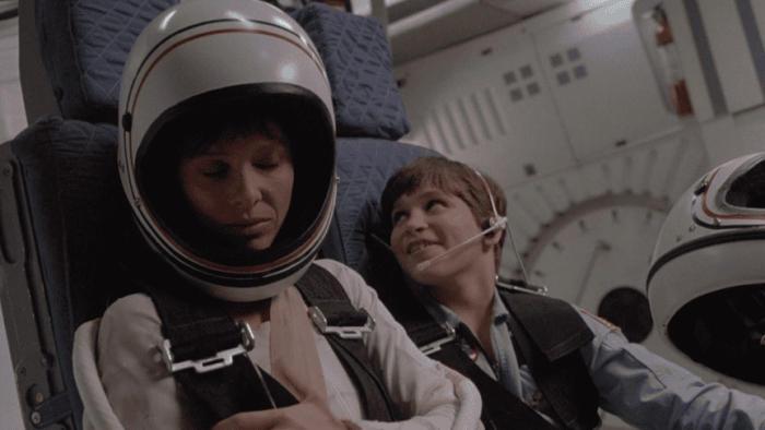SpaceCamp movie: Two people in spacesuits sit inside a spacecraft, with one person's eyes closed and the other person looking up.