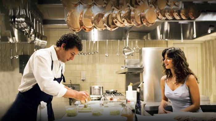 Two White adults with dark hair stand across a chef's table; the man at left wears chef's apron and looks preoccupied, while woman at right watches him