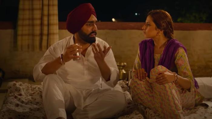 Saunkan Saunkne: While sitting on a mattress, an Indian man in a turban talks to an Indian woman