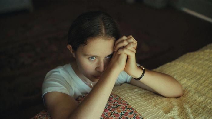 The Starling Girl Movie: A girl kneels next to a bed, her hands in a prayer position