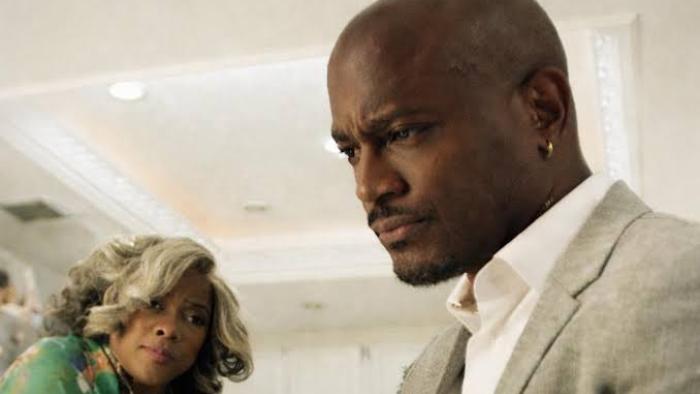 Stepfather Movie: Taye Diggs in a suit looking serious, Tamar Braxton in background watching, indoors.