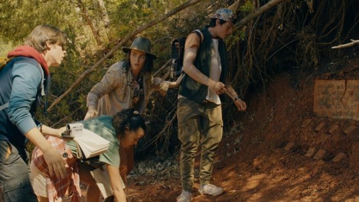 Four people stand at the mouth of a cave, one teen girl is reaching down to the ground. A teen boy wearing a backwards cap is studying a sign on the g