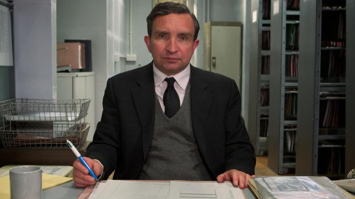 Still Life: Eddie Marsan wears a grey suit sitting at an office desk, a blank expression on his face