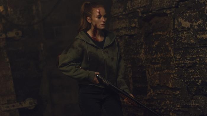 The Strangers: Chapter 3 Movie: Maya, injured and covered in blood, carries a shotgun into a kind of brick-walled cellar