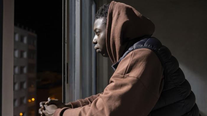 Street Flow 2 movie: Black French man in brown hoodie center looks left out window with apartment building in distance