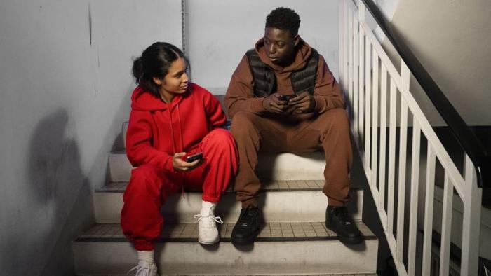 Street Flow 2 movie: Black French woman in red jumpsuit and hoodie left sits on stairwell next to Black French man in brown jumpsuit and hoodie right with railing on right
